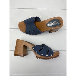 May include: Two vintage blue leather clogs with wooden heels. The clogs have a cutout design on the straps and silver-tone stud accents. The wooden soles are light brown, and the heels are approximately 3 inches high.