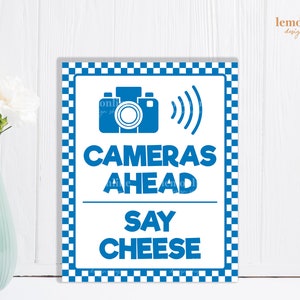 May include: Blue and white checkered sign with the text "Cameras Ahead Say Cheese" and a blue silhouette of a camera with a wavy line representing sound waves.