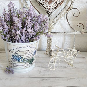 May include: White metal bucket with a lavender floral design and the text "Les Fleurs de Marseille Lavande" on the side. The bucket is filled with purple lavender flowers and is sitting on a white wooden surface. A white metal heart with a distressed finish and a white metal bicycle are also in the image.