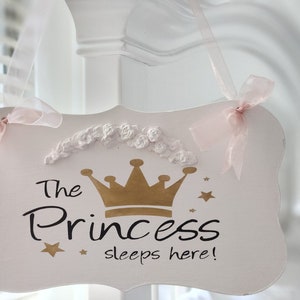 May include: White wooden sign with a gold crown and the text "The Princess sleeps here!"  The sign has a pink ribbon and a white floral trim.