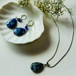 Op de afbeelding: Een set blauwe en zilveren oorbellen en een ketting met een blauwe stenen hanger. De oorbellen hebben een traanvormige steen en een kleine blauwe kraal. De ketting heeft een zilveren ketting en een traanvormige stenen hanger.