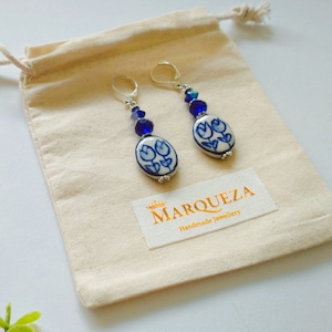 Puede incluir: Un par de pendientes de cerámica azul y blanca con un diseño floral, colgando de ganchos plateados. Los pendientes están adornados con cuentas de cristal azul y vienen en una pequeña bolsa de tela blanca con la etiqueta "MARQUEZA Joyería hecha a mano".
