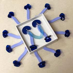 Puede incluir: Un par de pendientes de gradiente azul y transparente en una caja blanca. Los pendientes tienen forma de media luna y tienen un peque&ntilde;o amuleto de flor azul unido a la parte superior de cada pendiente.
