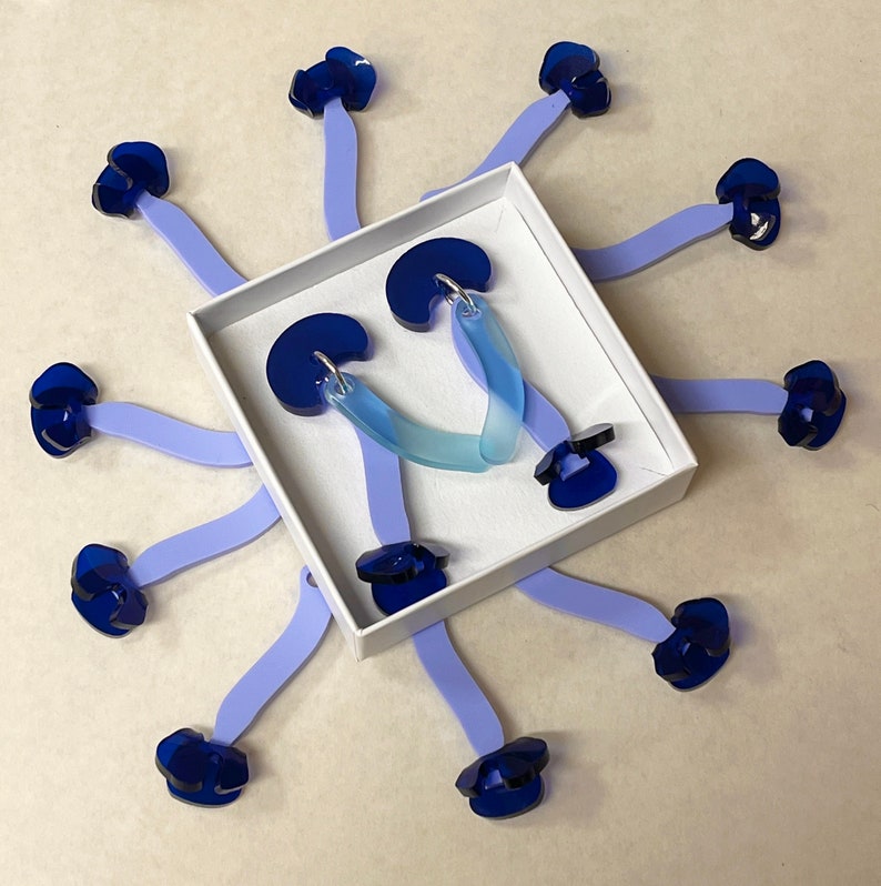 Puede incluir: Un par de pendientes de gradiente azul y transparente en una caja blanca. Los pendientes tienen forma de media luna y tienen un peque&ntilde;o amuleto de flor azul unido a la parte superior de cada pendiente.