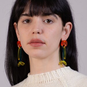 Puede incluir: Una persona con un su&eacute;ter blanco, cabello largo y oscuro y pendientes geom&eacute;tricos coloridos. Los pendientes est&aacute;n hechos de acr&iacute;lico naranja, amarillo y verde.