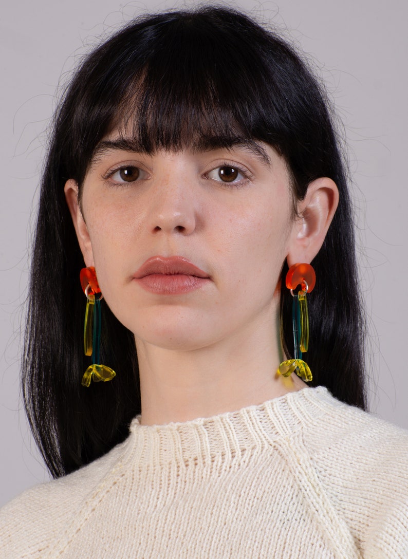 Puede incluir: Una persona con un su&eacute;ter blanco, cabello largo y oscuro y pendientes geom&eacute;tricos coloridos. Los pendientes est&aacute;n hechos de acr&iacute;lico naranja, amarillo y verde.