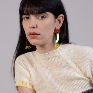 Puede incluir: Una mujer lleva un su&eacute;ter de punto blanco con ribetes amarillos y pendientes geom&eacute;tricos coloridos. Los pendientes est&aacute;n hechos de acr&iacute;lico y presentan una variedad de formas y colores, incluyendo rojo, naranja, amarillo, verde y azul.