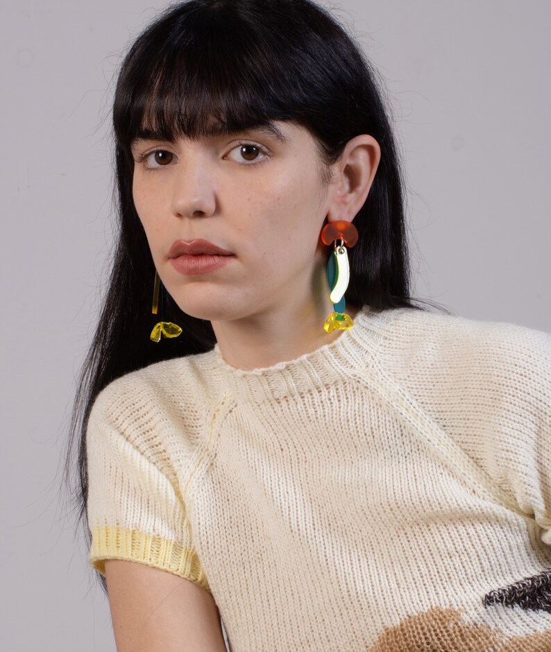 Puede incluir: Una mujer lleva un su&eacute;ter de punto blanco con ribetes amarillos y pendientes geom&eacute;tricos coloridos. Los pendientes est&aacute;n hechos de acr&iacute;lico y presentan una variedad de formas y colores, incluyendo rojo, naranja, amarillo, verde y azul.
