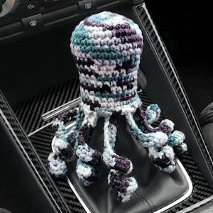 May include: A crochet octopus gear shift cover featuring a variegated yarn pattern in shades of dark purple, teal, light blue, and white. The octopus head sits atop the gear shifter, with eight curled tentacles hanging down around the base.