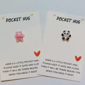 May include: Two white cards with the text "Pocket Hug" printed on them. Each card has a small, colourful charm attached. One charm is a pink bear and the other is a black and white panda. The text on the cards reads: "Here's a little pocket hug, please keep it safe and close, then it will be there beside you when you need it most."