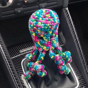 May include: A crocheted octopus gear shift cover featuring a vibrant, multi-coloured yarn pattern in shades of blue, pink, yellow, and green. The octopus has a rounded head and several dangling, curled tentacles that drape over the gear shifter.