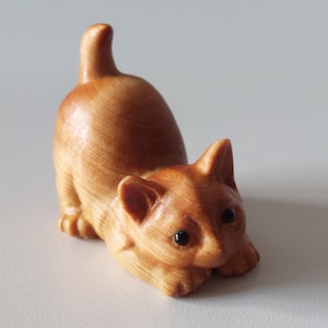 Op de afbeelding: Houten kattenbeeldje in een speelse pose, met uitgestrekte voorpoten en opgetrokken staart. De kat is gesneden uit lichtbruin hout, met zichtbare nerf en zwarte ogen. Decoratief item.