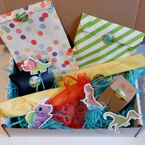 Pode incluir: Uma caixa de presente azul e branca cheia de papel de seda colorido, uma sacola de presente amarela e laranja, uma sacola de presente listrada verde e branca, uma pequena caixa marrom com a palavra "Dino" nela e vários adesivos de dinossauros.