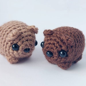 May include: Two crocheted stuffed animals, one brown and one light brown, with black button eyes. The animals are both sitting on a white surface.