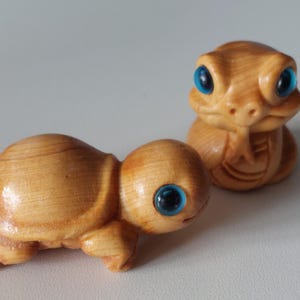 Puede incluir: Dos figuras de tortugas de madera con vetas de color marrón claro. Las tortugas tienen ojos azul brillante. Una tortuga está en posición de gateo y la otra está erguida. Las figuras son detalladas y parecen hechas a mano.