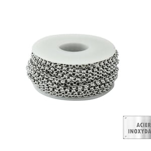 Könnte beinhalten: Eine Spule aus Edelstahlkette mit rundem Kettendesign. Die Kette ist um einen weißen Pappkern gewickelt. Ein kleines Metallschild mit dem Text "ACIER INOXYDABLE" befindet sich in der unteren rechten Ecke.