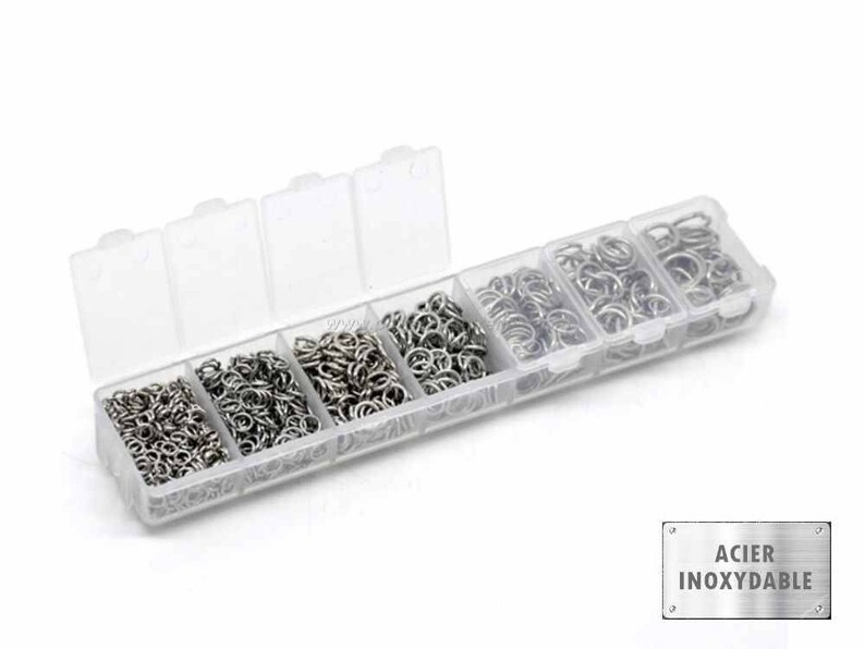May include: A clear plastic storage box filled with six compartments containing small silver jump rings. The box has a label that reads "ACIER INOXYDABLE".