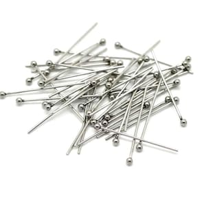 May include: A pile of silver-toned head pins with round heads. The pins are scattered on a white background.