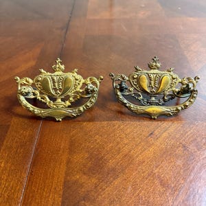 May include: Two antique brass drawer pulls with ornate, symmetrical designs. Each pull features a central, stylized vase shape with intricate scrollwork and a crown-like detail at the top. The pulls are set against a warm, wooden surface.