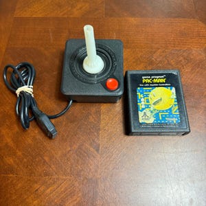 May include: A black Atari joystick controller with a white stick and a red button, connected to a black cord, alongside a rectangular Pac-Man game cartridge featuring a yellow character graphic and the text game program PAC-MAN.