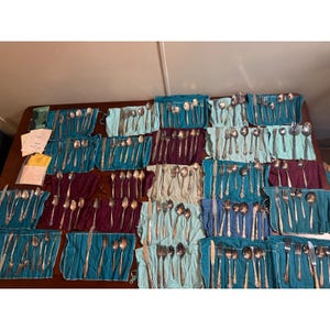 May include: A large collection of silverware sets, including forks, knives, and spoons, neatly arranged on a table. The silverware is wrapped in teal and burgundy fabric. Some sets are still in their original packaging.
