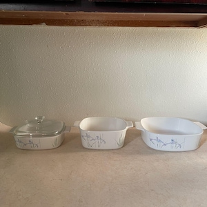 May include: A set of three white ceramic baking dishes with a floral design in blue and green. The set includes a square dish with a glass lid and two rectangular dishes. The dishes are arranged on a light-colored surface.
