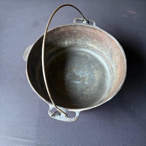 May include: A cast iron Dutch oven with a curved metal handle. The pot is dark brown with a slightly textured surface and two side handles. The handle arches over the top of the pot.