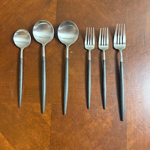 May include: A set of six stainless steel flatware pieces, including three spoons and three forks, arranged on a wooden surface. The spoons have rounded bowls and dark gray handles. The forks have three tines and matching dark gray handles. The flatware is designed for dining.