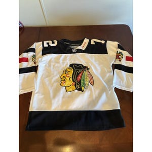 May include: White hockey jersey with black and red accents. Features the number 27 on the shoulders and a team logo on the chest. The jersey has a black collar and stripes on the sleeves.