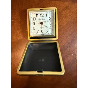 May include: A vintage gold-tone travel alarm clock with a square face and a hinged cover. The clock face is white with black numerals and hands, and the text "Seth Thomas" is visible. The alarm clock is open, revealing a black interior.