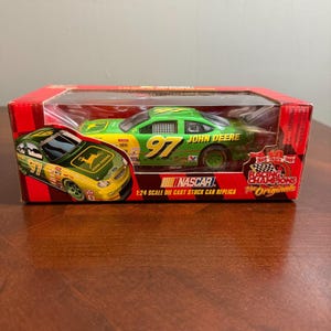 May include: A green and yellow John Deere NASCAR die-cast stock car replica in a red box. The car features the number 97 and the John Deere logo. The box reads "Racing Champions The Originals".