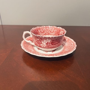 May include: A red and white floral patterned teacup and saucer set. The teacup has a scalloped rim and a curved handle. The saucer is round and matches the teacup's design. The set is made of ceramic.