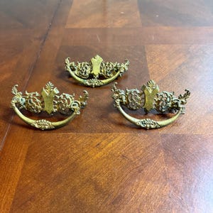 May include: Three antique brass drawer pulls with ornate, curved handles and decorative centerpieces. The pulls have a vintage aesthetic, with intricate floral and scrollwork details. The brass has a slightly aged patina, adding to their classic charm.
