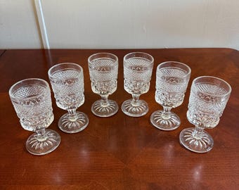 May include: Six clear, vintage-style stemmed glasses with a diamond-cut pattern. The glasses have a round base and a short stem. The glasses are arranged in a row on a dark wooden surface.