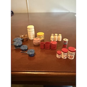 May include: A collection of miniature kitchen items, including blue speckled pots, milk bottles, and various canisters labeled "Milk", "Cookies", "Sugar", and "Flour". A small Pepsi can is also present. The items are arranged on a brown surface.