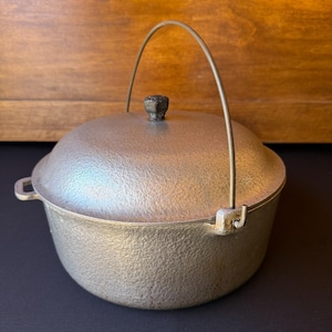 May include: A silver-colored, cast-iron cooking pot with a lid and a handle. The pot has a textured surface and a small knob on the lid. The handle is arched and attached to the sides of the pot. The pot is sitting on a dark surface.