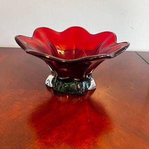 May include: A red glass bowl with a scalloped rim and a green glass base. The bowl is sitting on a wooden surface.