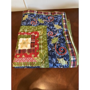 May include: A folded Christmas-themed quilt with a patchwork design. The quilt features a combination of plaid, floral, and patterned fabrics in red, green, blue, and gold. The edges are trimmed with a green and red plaid fabric.