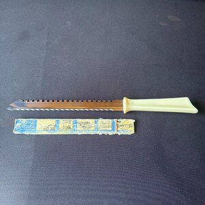 May include: A serrated bread knife with a cream-colored handle and a partially visible blue and white paper sleeve. The knife blade is stainless steel.