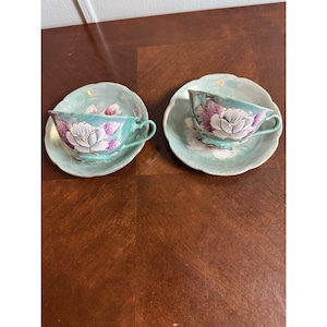 May include: Two matching teacups and saucers. The teacups and saucers are a light teal color with a floral design featuring white and purple flowers. The teacups have a curved handle and the saucers have a scalloped edge.