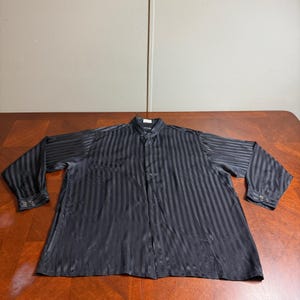 May include: A long-sleeved button-down shirt in a solid black color featuring a subtle vertical striped pattern throughout the fabric. The shirt has a standard collar, a front button placket, and cuffs on each sleeve that are secured with buttons.