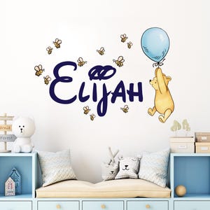 May include: A wall decal featuring the name "Elijah" in navy blue script, surrounded by cartoon bees. A yellow bear holds a light blue balloon. The decal is designed for a child's room.