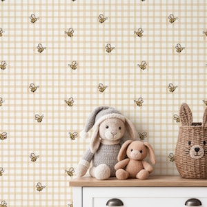 May include: A whimsical scene featuring a beige and white checkered wallpaper adorned with small bee illustrations. A gray bunny plush toy with a matching hat, a small brown bunny plush, and a woven basket shaped like a bunny are on a wooden surface.
