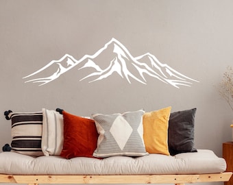 May include: A white mountain range silhouette wall decal above a light beige couch with black, white, and brown striped throw pillows, a rust colored velvet pillow, a black and white geometric pillow, and a mustard yellow pillow.