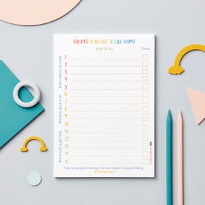 Könnte beinhalten: Ein weißer Notizblock mit dem Text "TODAY'S TO DO LIST TO LIVE HAPPY" in Regenbogenfarben. Der Notizblock hat eine Liste von 15 Elementen mit Kontrollkästchen. Zwei Bleistifte und dekorative Formen liegen auf der grauen Oberfläche.