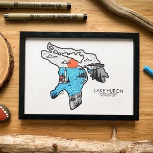 May include: A framed illustration of Lake Huron, featuring a lighthouse, sun, and water. The artwork is in a black frame and includes the text "Lake Huron, Ontario Canada, Michigan USA". The image is surrounded by art supplies and natural elements.