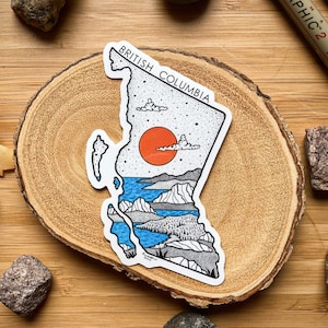 May include: A sticker in the shape of British Columbia, with a landscape design including a red sun, blue water, and mountains. The text "BRITISH COLUMBIA" is at the top. The sticker is on a wooden slice, surrounded by rocks and leaves.