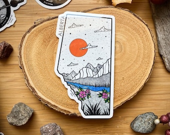 May include: A sticker of the province of Alberta, Canada. The sticker is white with black outlines and features a mountain range, a river, a sun, and clouds. The text "ALBERTA" is printed in black at the top of the sticker.
