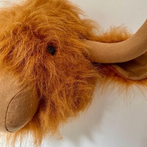 Peut inclure: Une t&ecirc;te de vache Highland en peluche marron et beige avec un pelage moelleux et hirsute. La t&ecirc;te est mont&eacute;e sur un mur blanc.