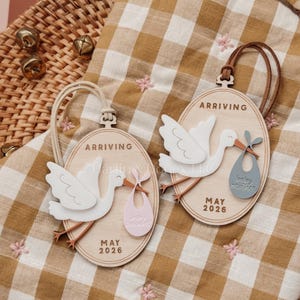 Può includere: Due ornamenti ovali in legno con una cicogna che trasporta un bambino. Gli ornamenti hanno la scritta "ARRIVING" in alto e "MAY 2026" in basso. Un ornamento ha un bavaglino rosa, l'altro un bavaglino blu. Gli ornamenti sono su un tessuto a scacchi marrone e bianco.